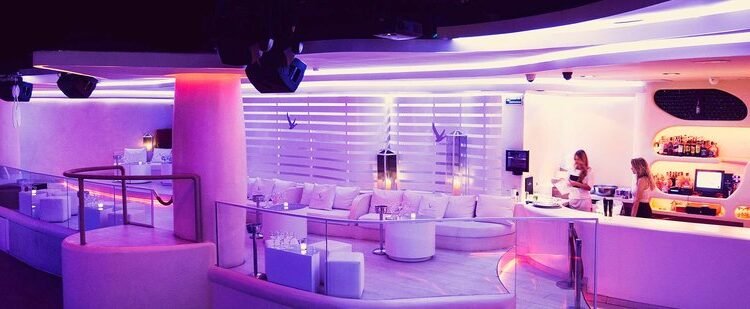 Interior de la discoteca Pacha Barcelona: zona VIP con mesas iluminadas, sofás blancos y luces rojas y púrpuras. El ambiente es nocturno, con decoración moderna y elegante, y la pista de baile se ve al fondo bajo focos y efectos de luz