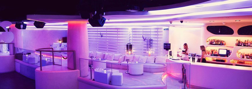 Interior de la discoteca Pacha Barcelona: zona VIP con mesas iluminadas, sofás blancos y luces rojas y púrpuras. El ambiente es nocturno, con decoración moderna y elegante, y la pista de baile se ve al fondo bajo focos y efectos de luz